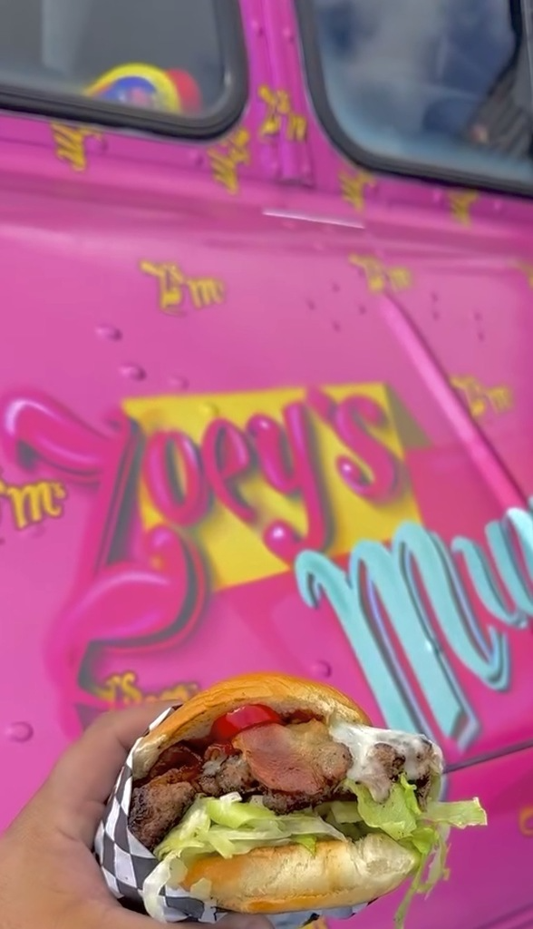 A bacon burger held in front of the pink Zoey's Munchies truck